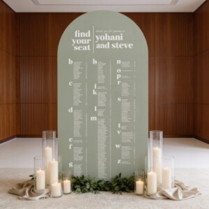 Sage green arched seating chart backdrop for Yohani and Steve's wedding, surrounded by candles and greenery